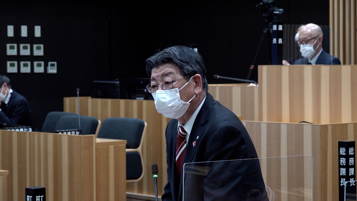 議場で、黒いスーツに赤いネクタイを締めた佐藤仁町長が立ち、マイクの前で発言しており、背後には議長席や撮影用のカメラも見える議会中の写真