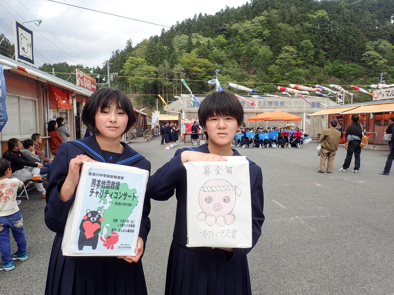 さんさん商店街の前で、熊本地震被災地支援チャリティーコンサートのポスターを貼った募金箱とオクトパス君のイラストが描かれた募金箱を手に持った女子中学生が並んで立っている写真