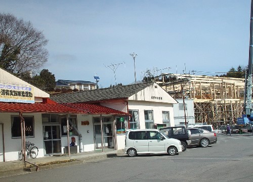 日差しの明るい広場のような駐車場の手前に、平屋の建物が並んでおり、その奥の場所に木造の建物の骨組みを組んでいる建設途中の様子が見える歌津公民館の写真