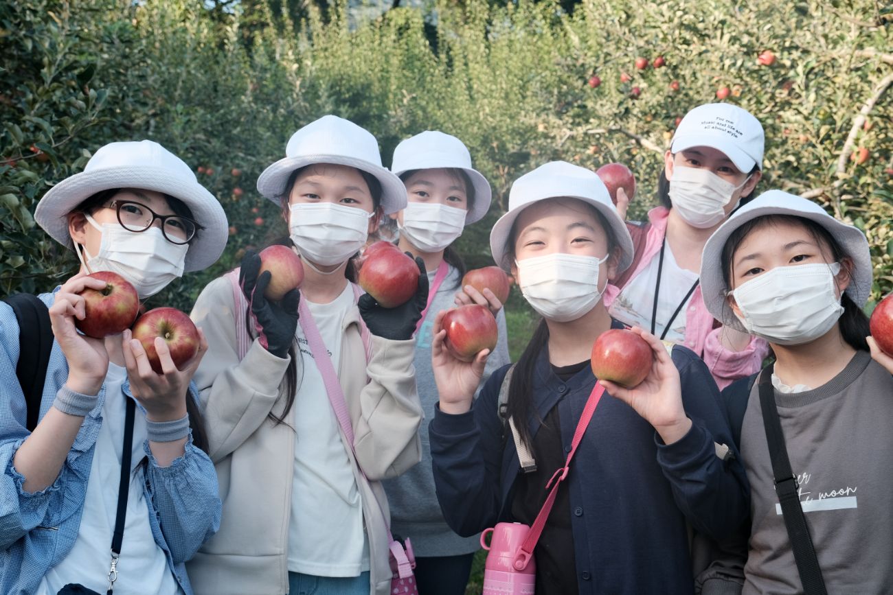 りんご園で白い帽子とマスクを着けた6人の児童が両手で赤いりんごを持ち笑顔でカメラに向かって並んでいる様子の写真