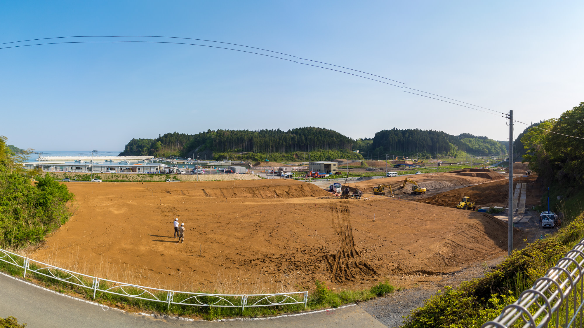 やや高い位置から見下ろす定点カメラによる昼間の風景で広く平らに整地された赤茶けた土の空き地の中央付近に、白いヘルメットをかぶった作業服姿の人物が2人、向かい合って立っている様子の写真