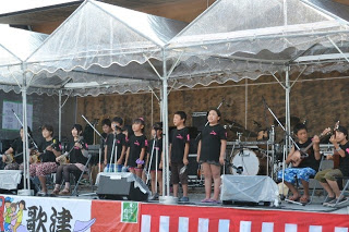 黒いTシャツを着た子どもたちが、マイクの前に立って合唱を行い、横並びに三味線などの楽器を演奏している「ポストくんおかえりコンサート」の様子を写した写真