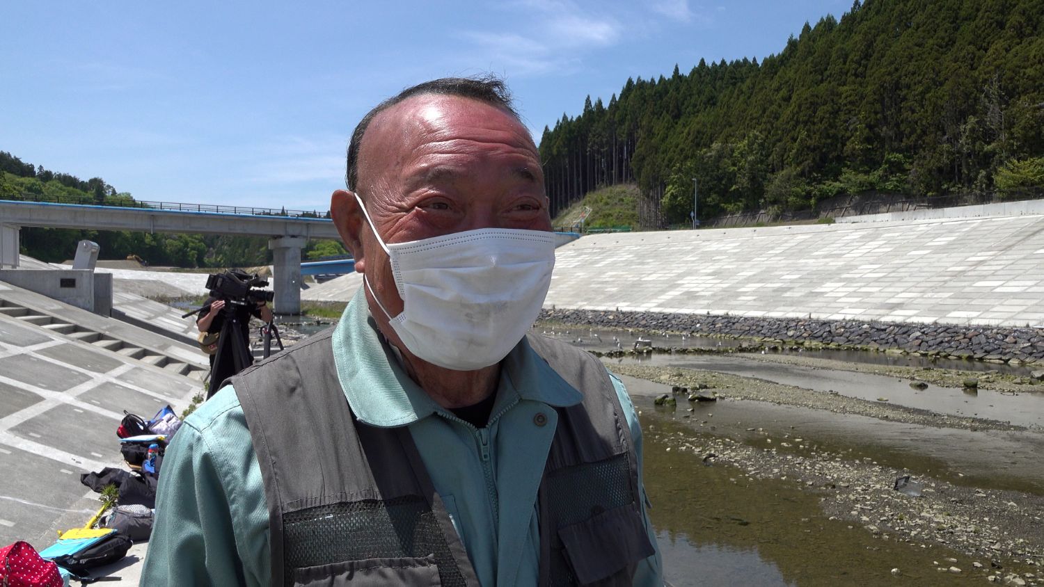 河川の護岸の前でマスクをしたベスト姿の渡邊さんが笑顔で立っている様子をアップで撮影した写真
