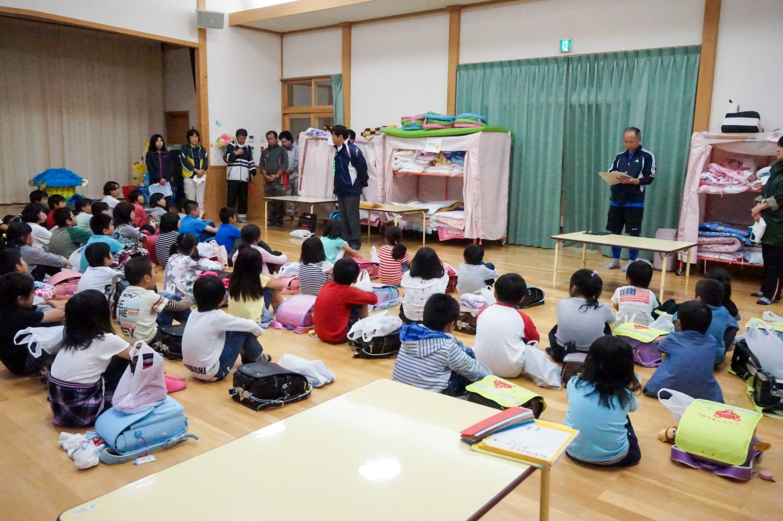 多くの子どもたちが室内で床に座り、大人が前に立って話をしているのを静かに聞いている様子の写真