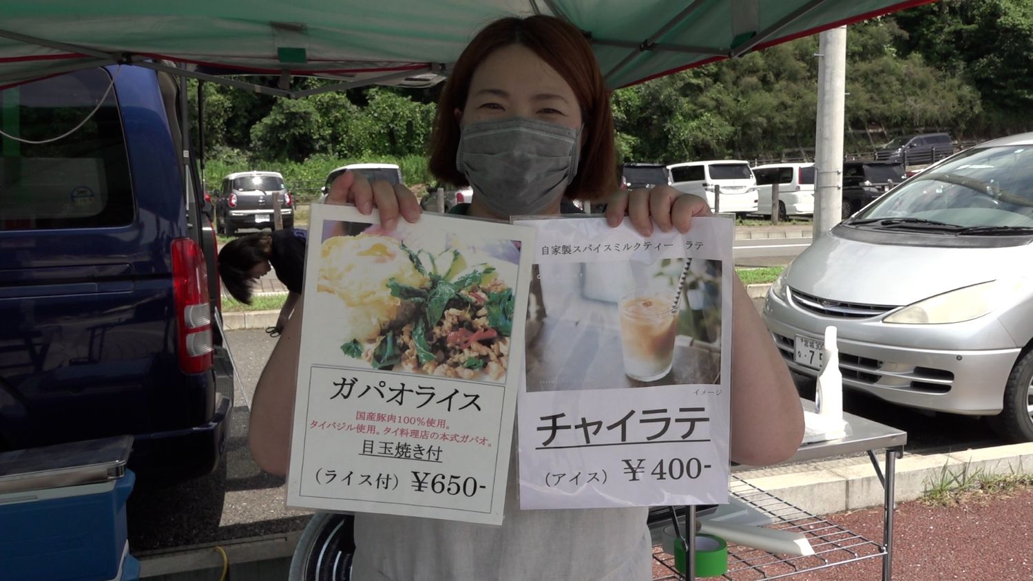 青と銀の車が並んでいる前でマスクを着けた女性が屋台のテントの下でガパオライスとチャイラテのメニューを両手に持っている写真