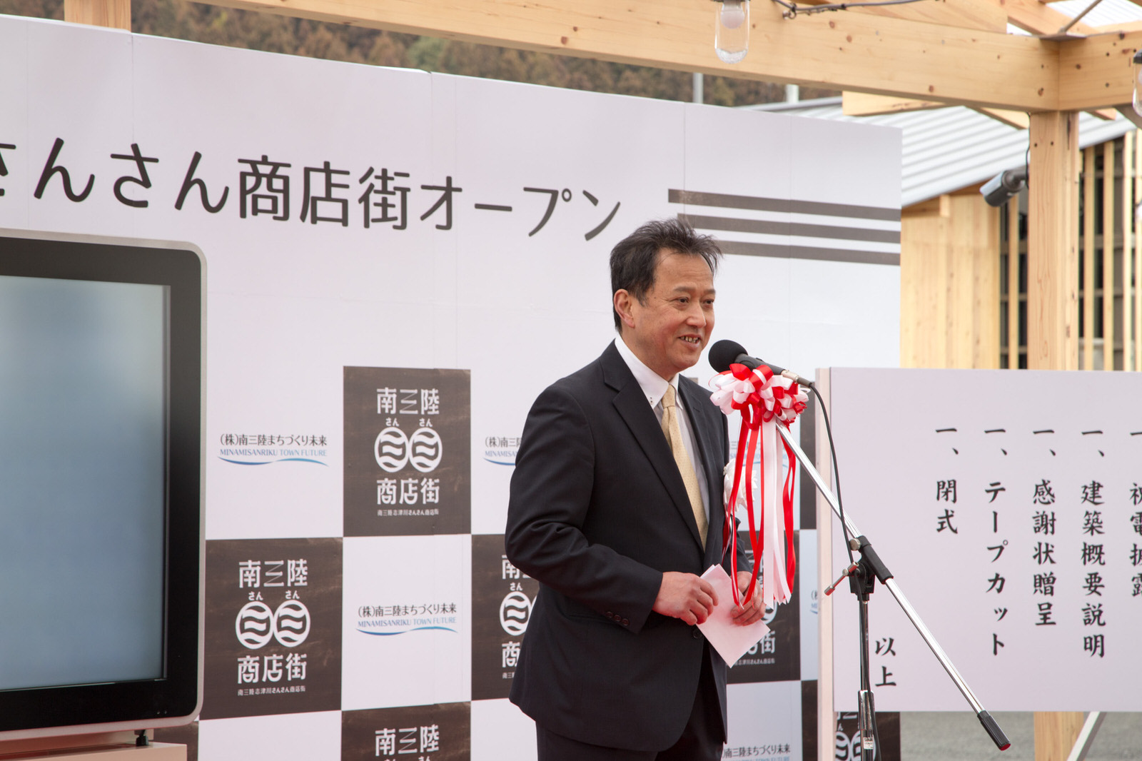 スーツ姿の三浦社長が「南三陸志津川さんさん商店街オープン」と書かれた看板や式次第が掲示されたマイクスタンドの前に立ち、開会の挨拶を行っている様子の写真