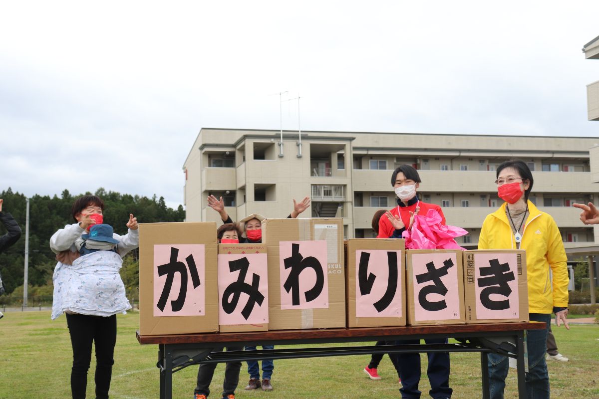 「かみわりざき」と一文字ずつ書かれた紙が貼られた段ボール箱が6個並び、その後ろでスタッフや参加者が声援や応援をしている様子を写した写真