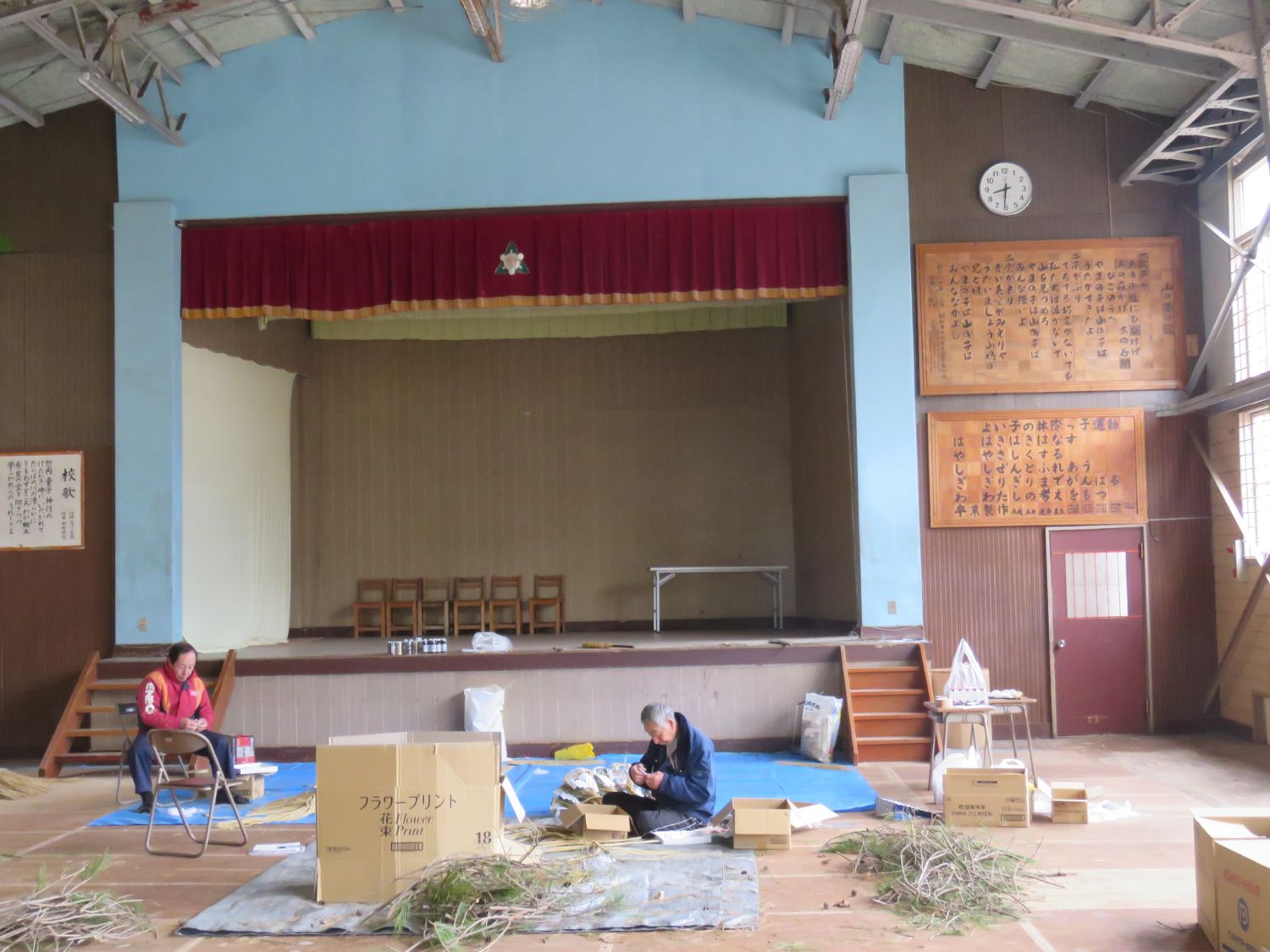 パイプ椅子に座りしめ縄を編んでいる山内さんとあぐらを組んで床に座りしめ縄を編んでいる佐藤さんの様子を遠くから撮影した写真