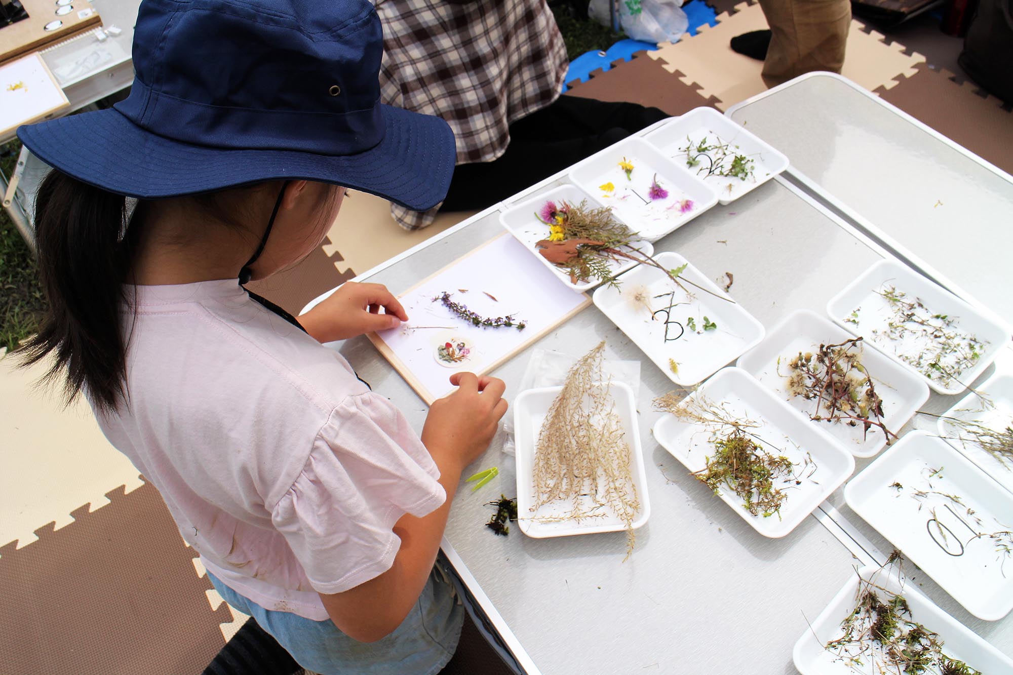 帽子をかぶった子どもが白いトレーに並べた草花を使って、紙の上で自然素材のバッジを作るワークショップ体験の様子の写真