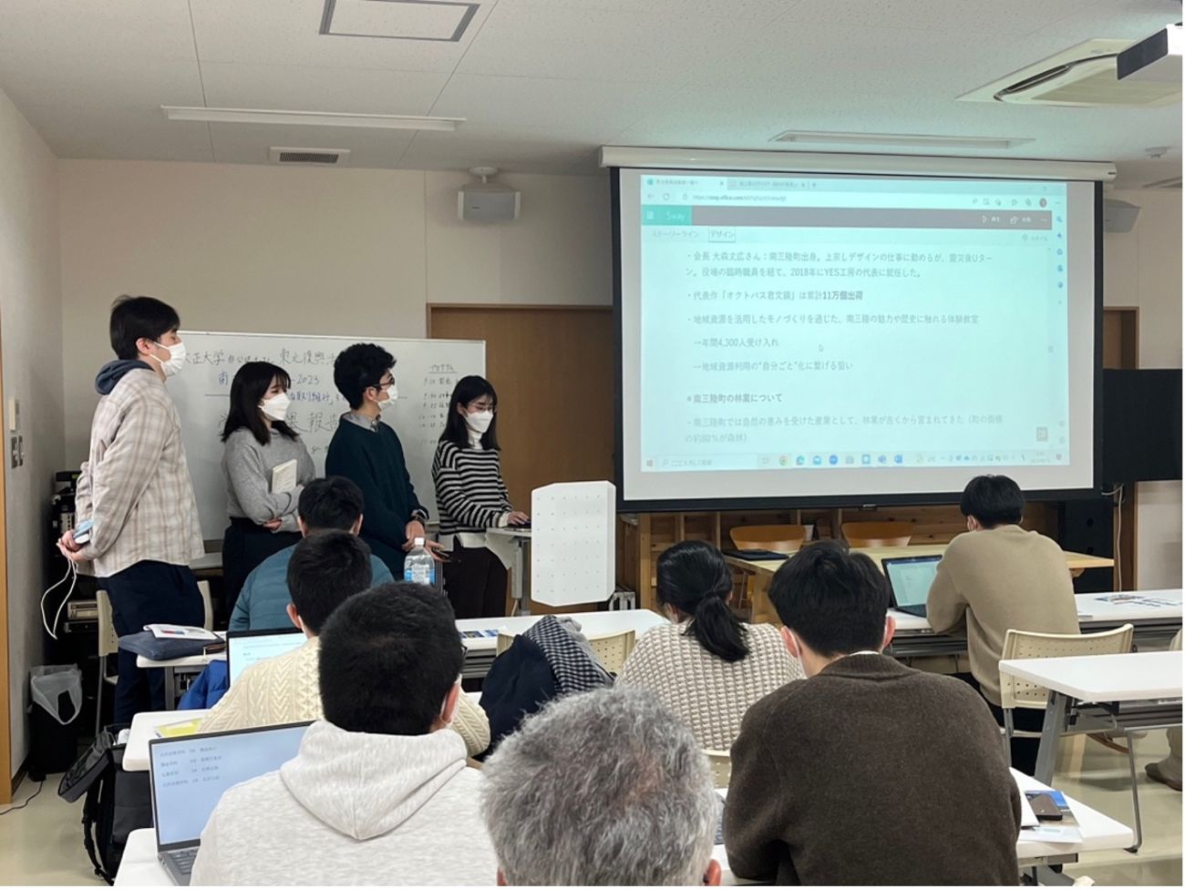 室内の会議室で数人の若者が前に立ち、ホワイトボードとスクリーンを使って他の参加者に向けて発表しており、スライドには地域活動や復興に関する説明が書かれている写真