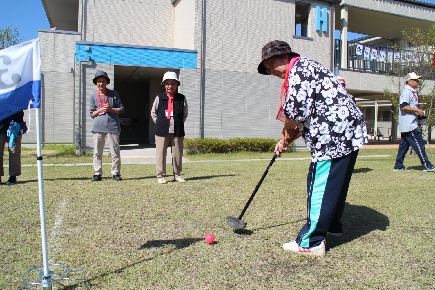 数人が見守る中、立っている旗を目掛けて女性が赤いボールを打とうとしているグラウンドゴルフの様子の写真
