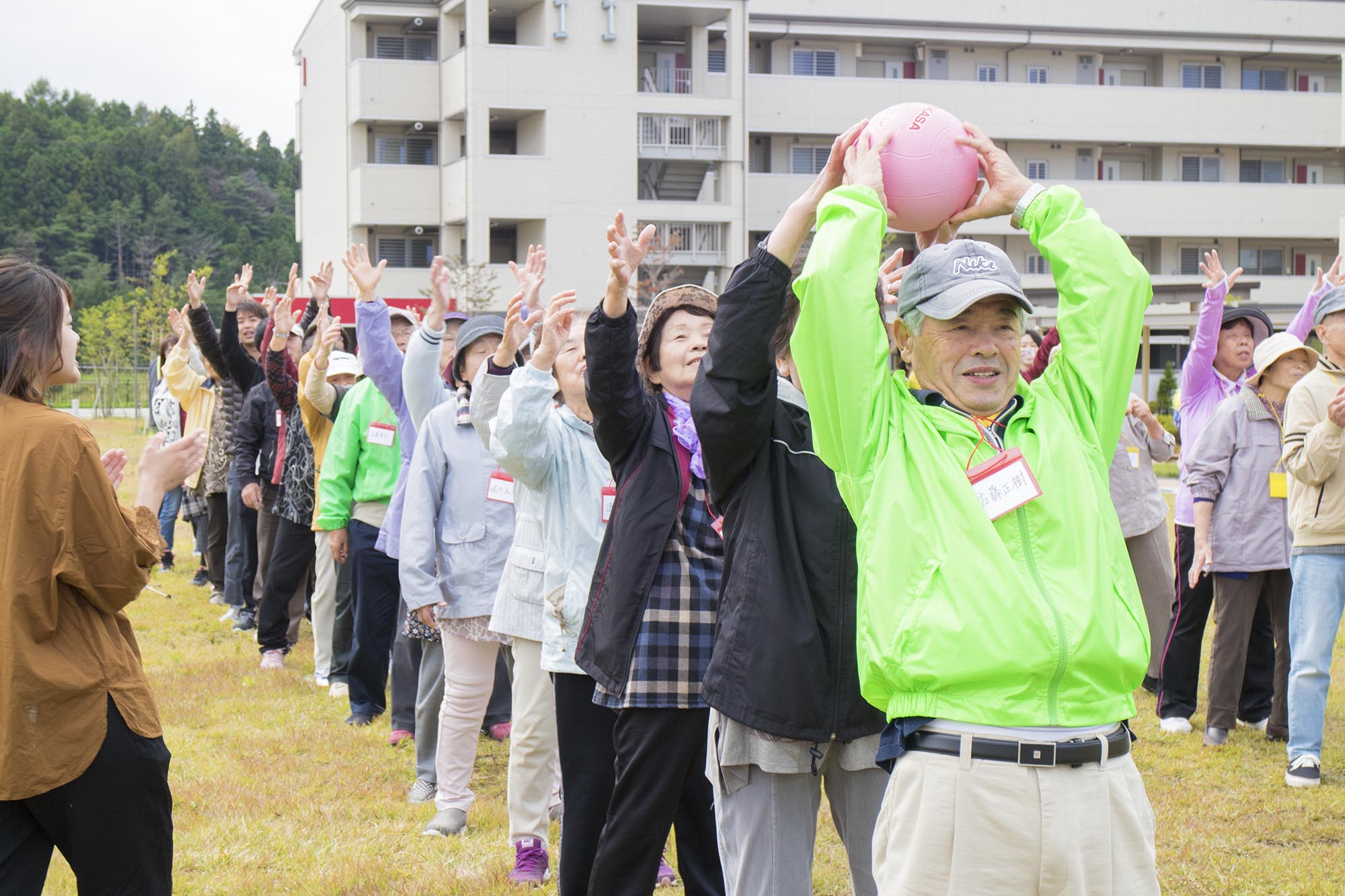 縦一列に並び、先頭の男性がピンクのボールを頭の上で持ち、後ろの人たちが両手を挙げて次に渡すリレー形式の競技をしている写真