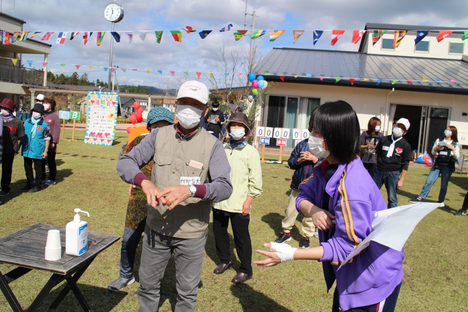 芝生の広場に机が設置され消毒薬が置かれ参加者が一列に並び手を消毒しているのを関係者が見守っている様子の写真