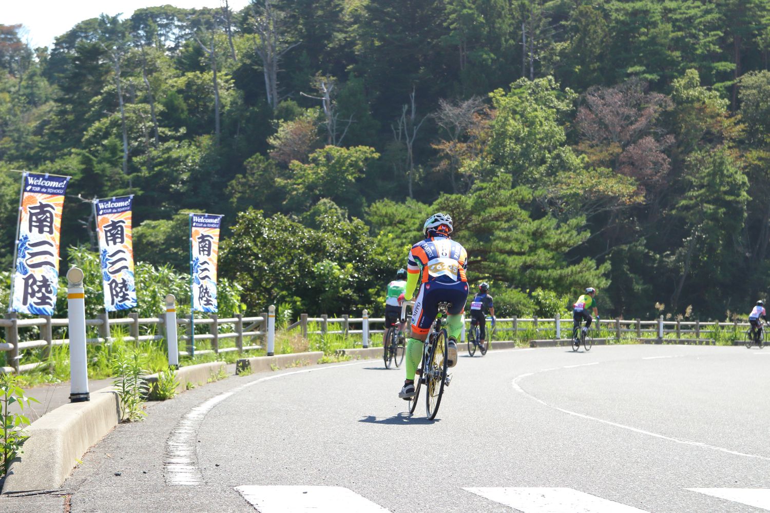 等間隔に「南三陸」と書かれたのぼり旗が立っている道路を、頭にヘルメットを被ったライダーの方々が縦一列に並んで走行している様子を後方から撮影した写真