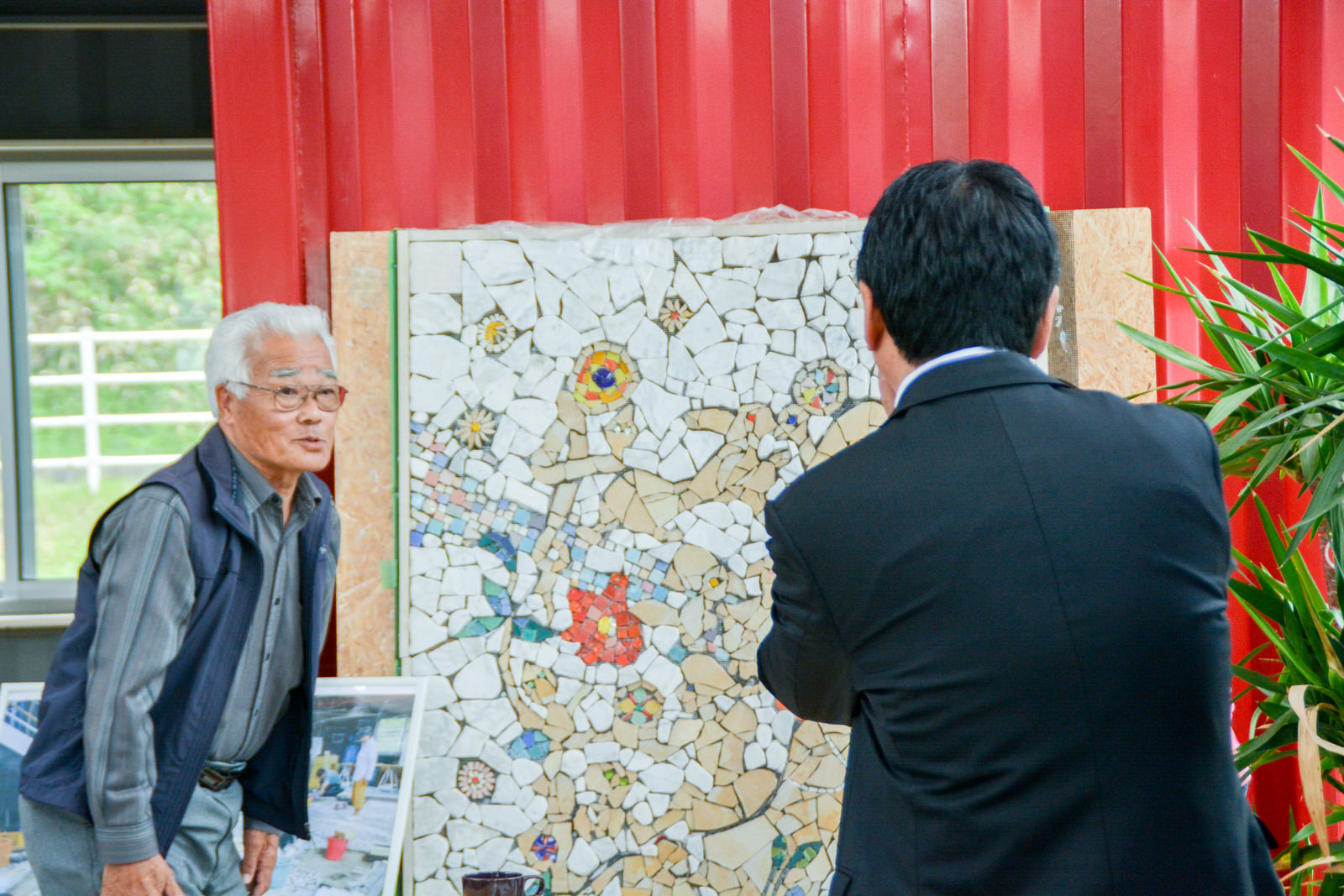 モザイクアートのモニュメントの横に白髪の男性が立っており、モニュメントを見ている町長の後ろ姿を写した写真