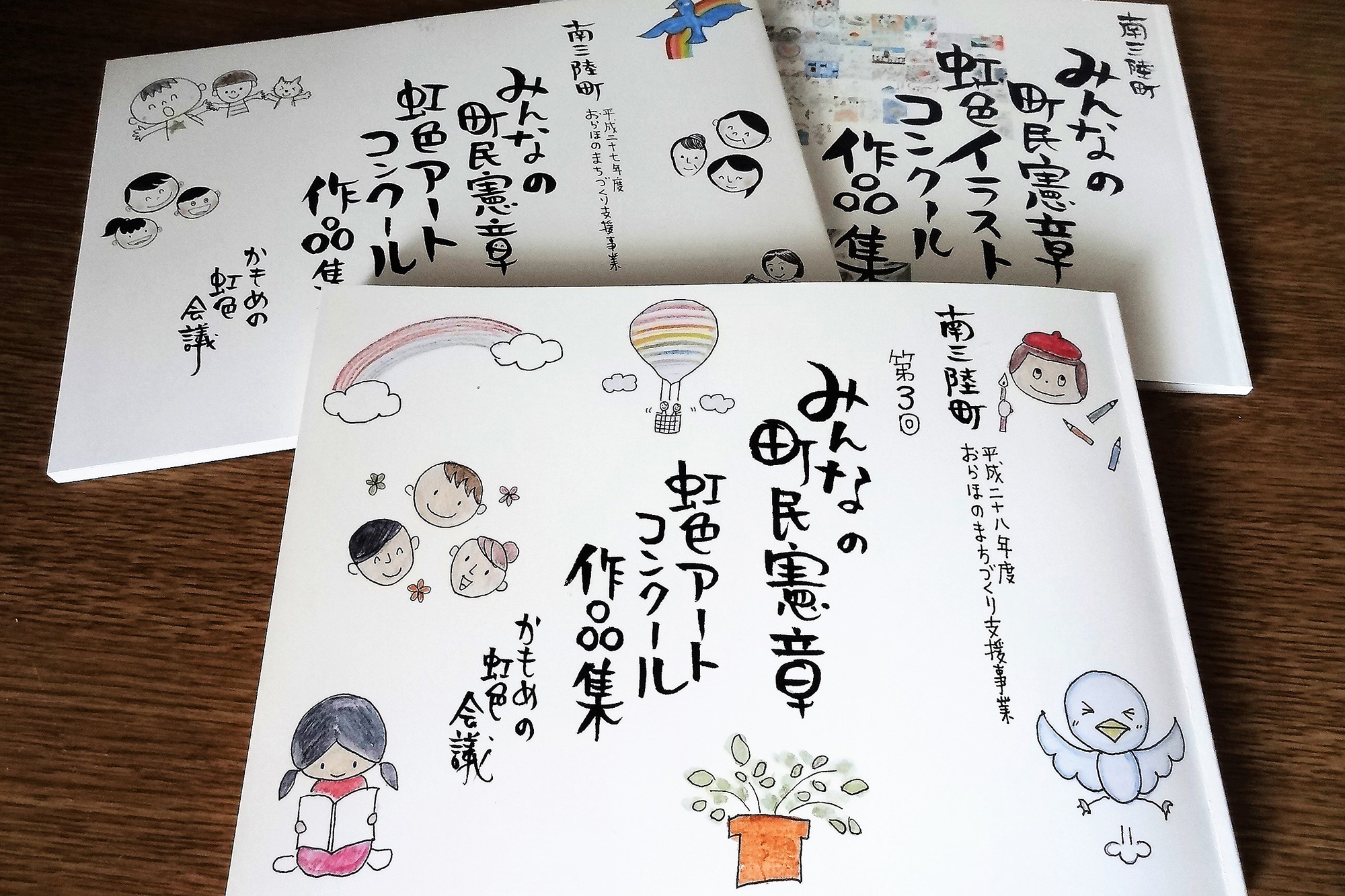 『みんなの町民憲章 虹色アートコンクール作品集』と書かれた冊子が3冊重なって並べられていて、表紙には手書き風の文字と虹や気球、笑顔の人々、植物などのイラストが描かれている写真
