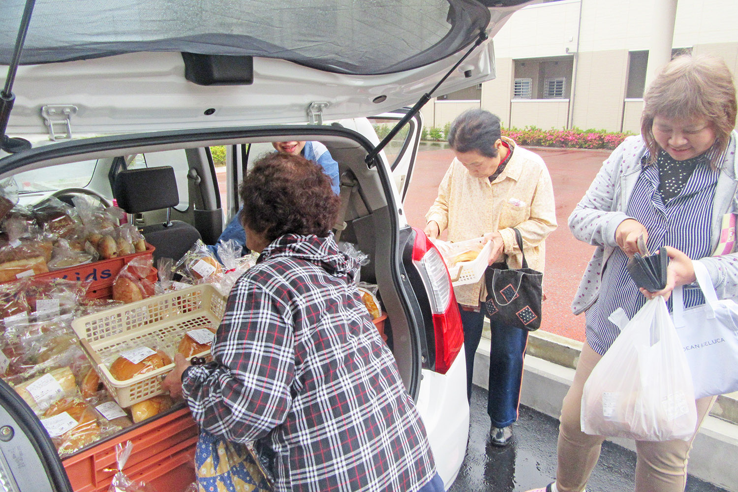 移動販売車の後ろに並べられた沢山のパンを数人の女性がかごに入れたり財布を取り出したりして購入している写真