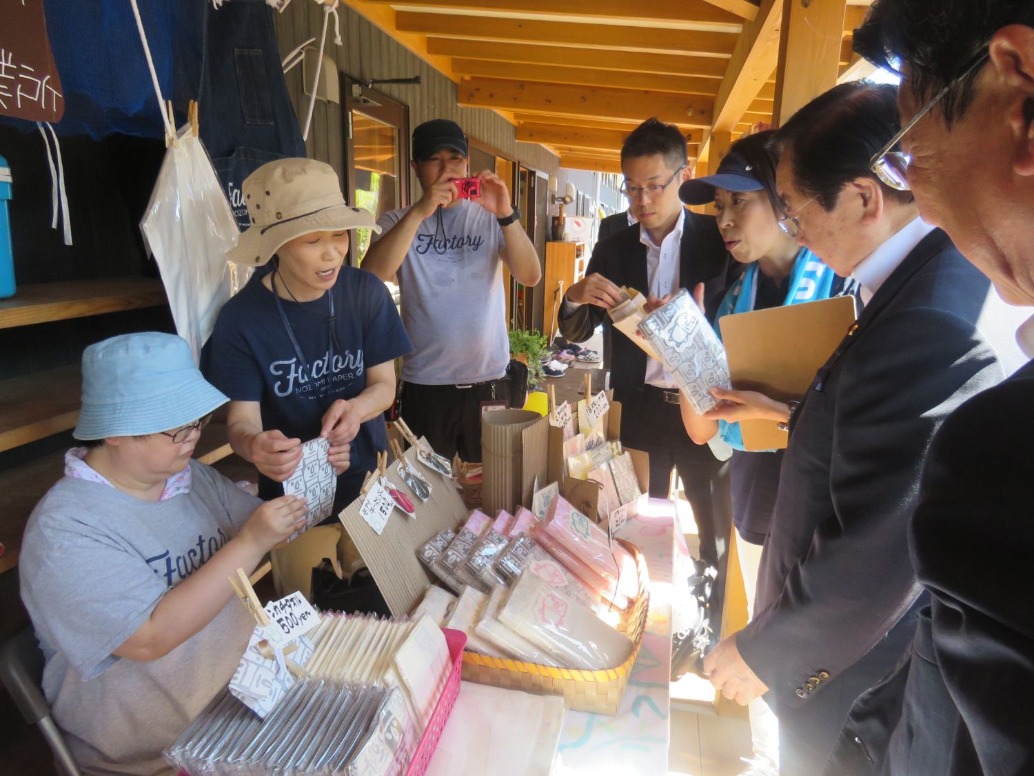 屋根付きの屋外通路で、「Factory」と書かれたTシャツを着たスタッフが、テーブルに並べた布製品や手作り雑貨をスーツ姿の来場者たちに説明しながら販売しており、商品には丁寧に値札が付けられ、和やかな交流が行われている写真