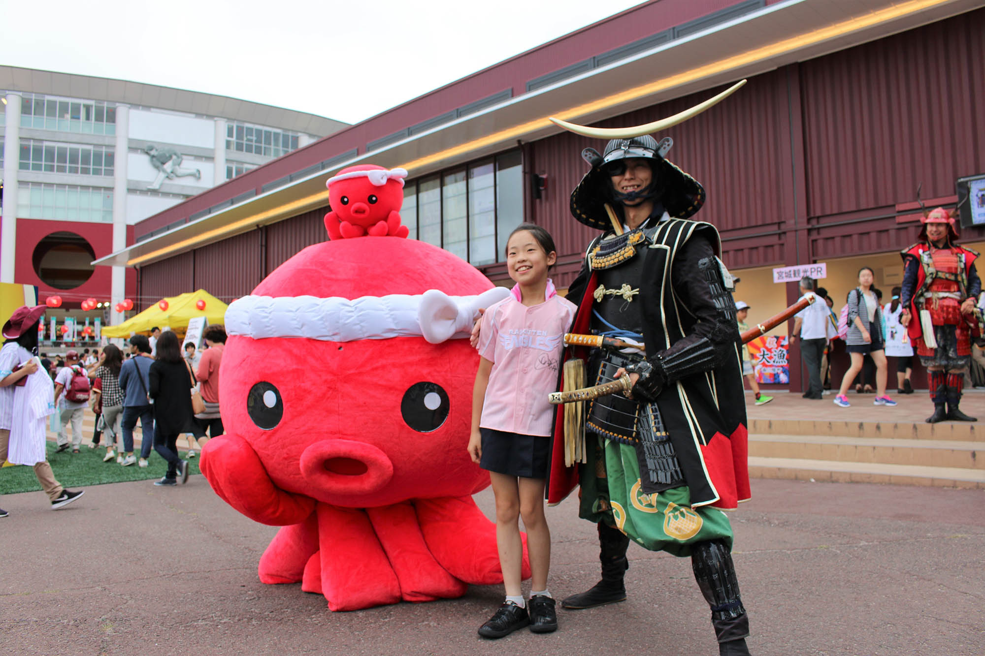 巨大な赤いタコの「オクトパス君」が頭に小さな「オクトパス君」を頭に乗せ、笑顔の女の子と黒い甲冑を着た侍の格好をした男性と一緒に野外イベント会場でポーズをとっている写真