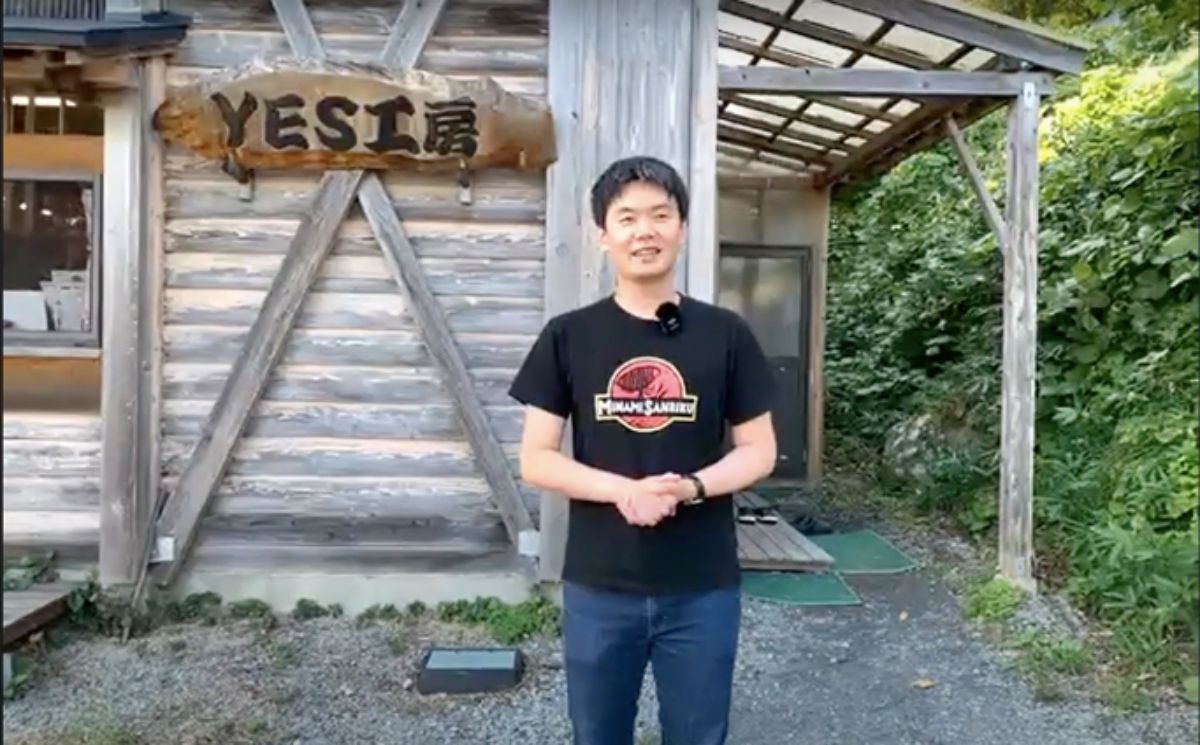 建物の右側には緑の植物が生い茂り、木の板でできた壁に「YES工房」と書かれた看板が掛かっている木造の建物の前で、黒いTシャツを着た男性が立っている写真