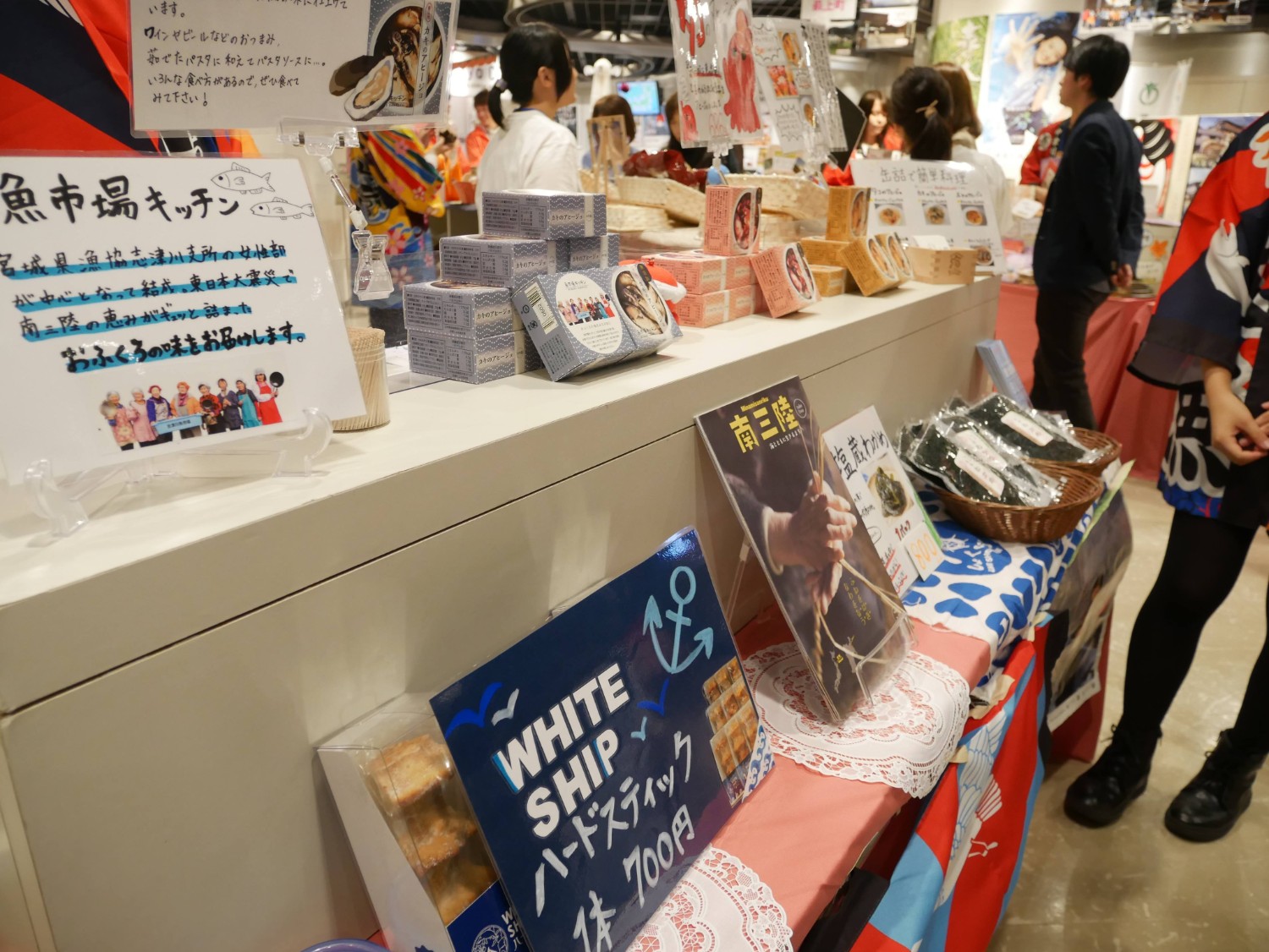 法被を着た販売員が接客する屋内の市場の一角で、カウンターの上に商品の説明や値段が書かれたポップやパッケージが並び、来場者が商品を見ている様子の写真