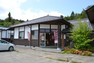 白い壁と黒い瓦屋根の木造の建物で、建物の前には赤い旗と看板が立ち、周囲には緑の木々や草花の植栽が植えられている南三陸森林組合の外観写真