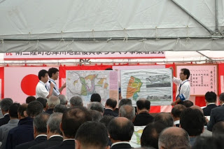 壇上に立つ担当者たちが地図や計画図を持ち上げて、出席者に向けて復興事業の説明をしている写真