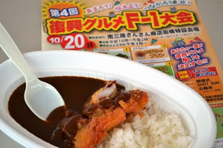 白い容器に盛られたさんさんタコカレーがテーブルに置かれ、その奥に「第4回 復興グルメF-1大会」と書かれたカラフルなチラシが置かれている写真