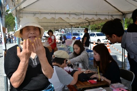 白いテントの下で、両手の爪に黄色のネイルをしてもらった女性が、嬉しそうにカメラに向かって手を見せている様子の写真