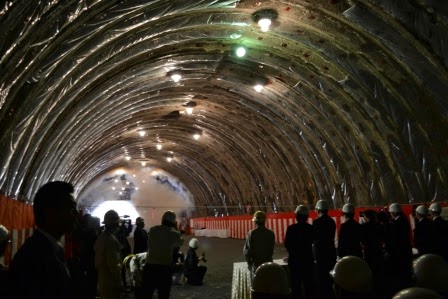 壁を貫通してもくもくとした煙が残り、太陽の光が差し込んで天井の照明も灯されて明るくなったトンネル内で、ヘルメットを着用した人々や、カメラを構えていた左側に座っている人たちも後ろを振り返っている貫通後の様子の写真