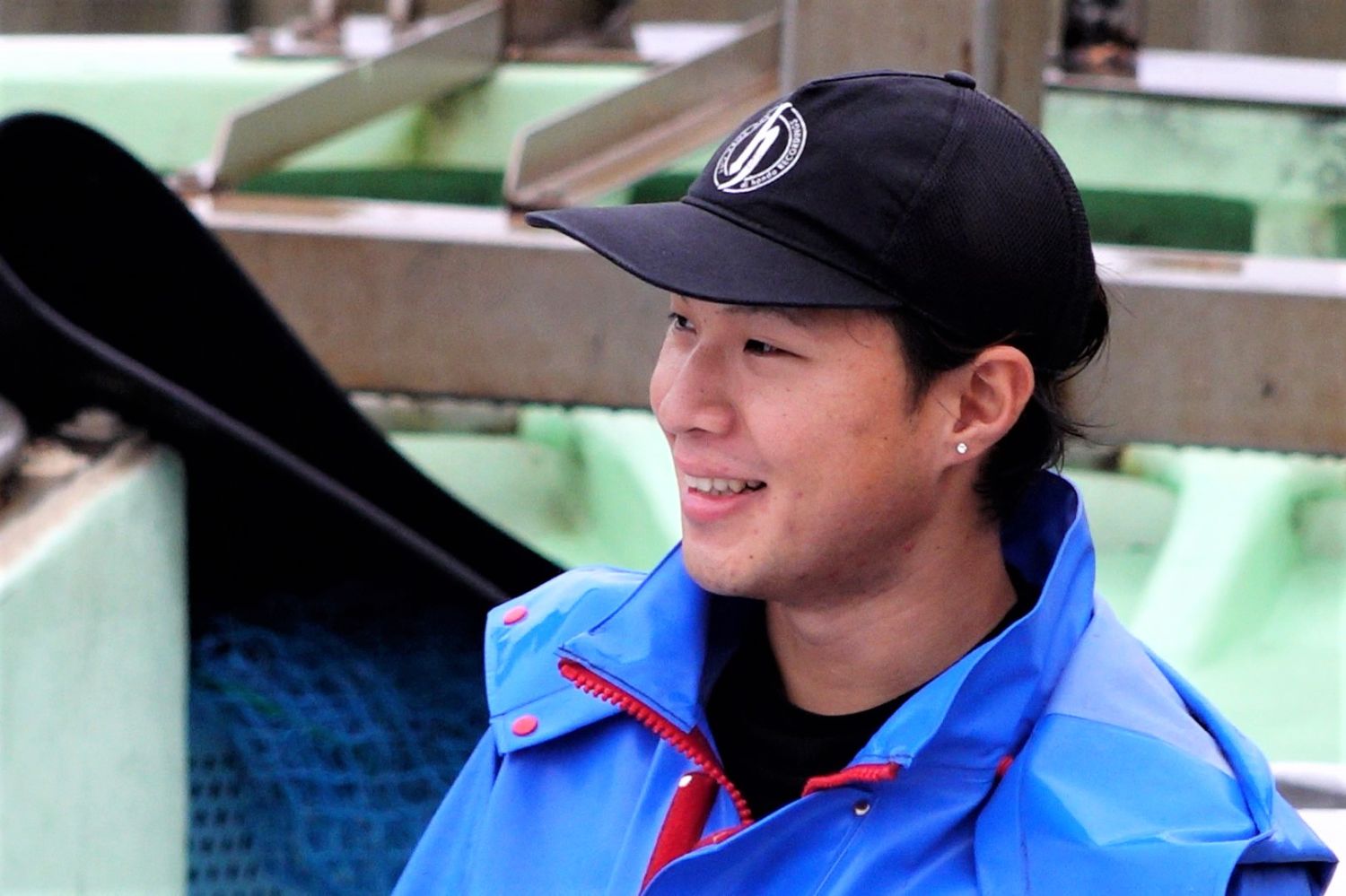 青い合羽を着て黒い帽子をかぶった吉岡さんが船上で笑顔を浮かべており、後方には漁で使用される機材が見える、作業の合間の表情を捉えた写真