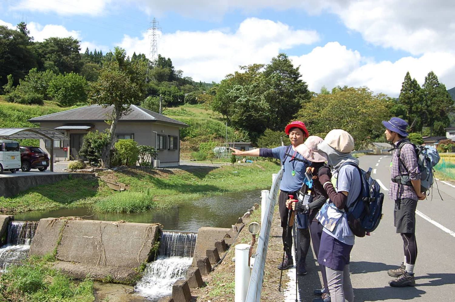 田舎道のそばで、小川を見ながらガイドの話に耳を傾けているハイキング中の人々の様子の写真
