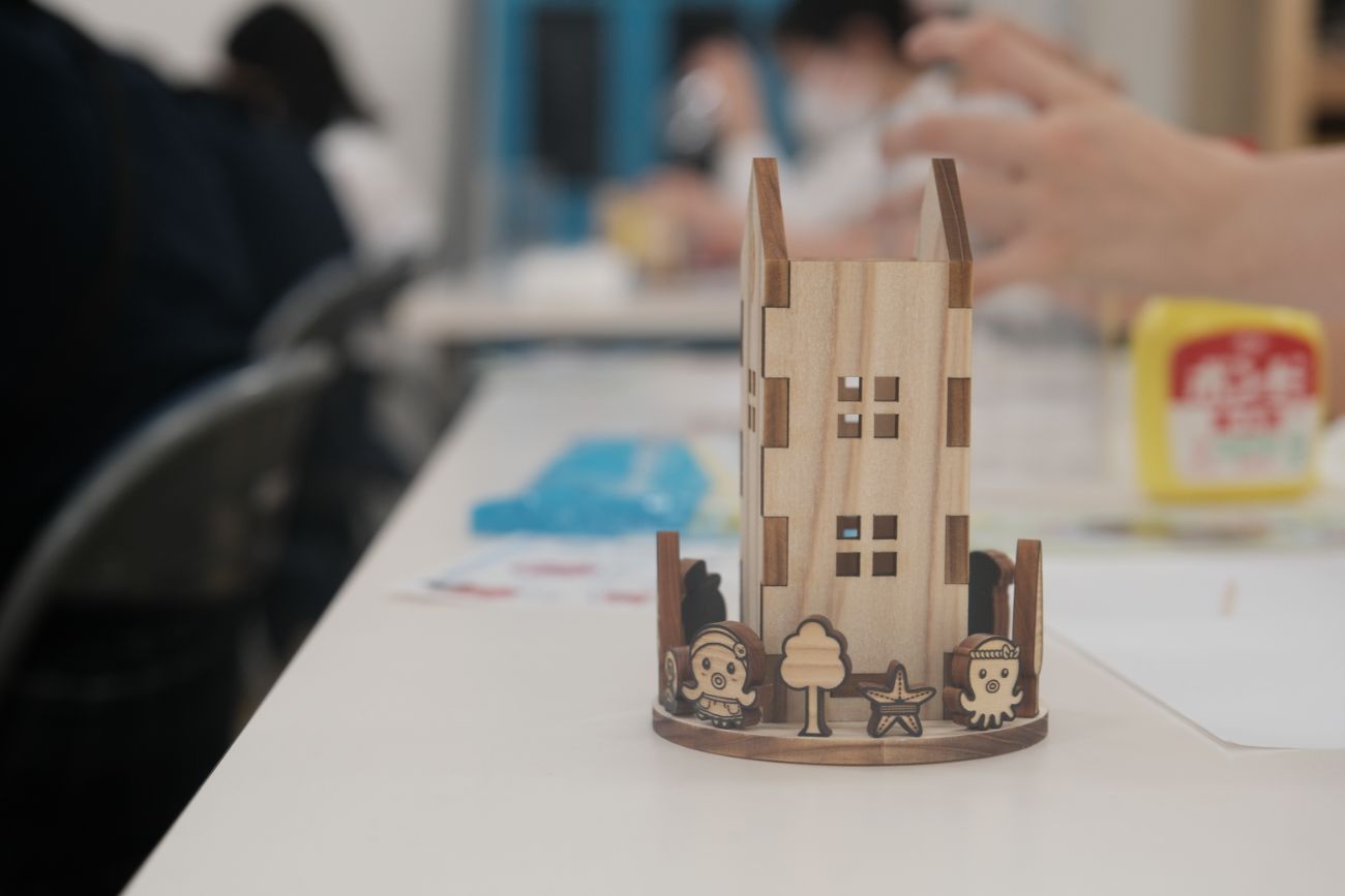 木製の高い建物型ペンスタンドの完成品が机の上に置かれており、土台には木やタコのキャラクター、星形の飾りなどが貼られている写真
