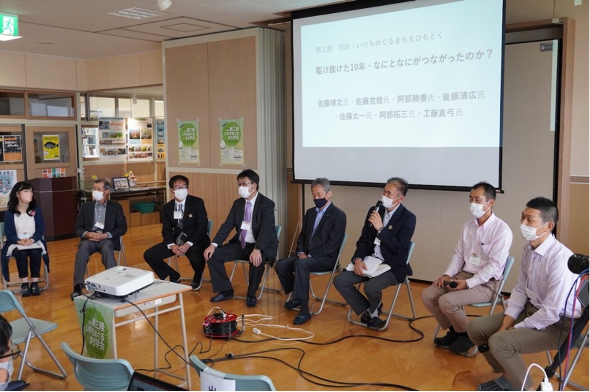 室内の会場で、プロジェクターにスライドを映しながら、スーツや名札をつけた8人の登壇者が椅子で丸く並んで座り、マイクを使って観客に向けてパネルディスカッションをしている様子の写真