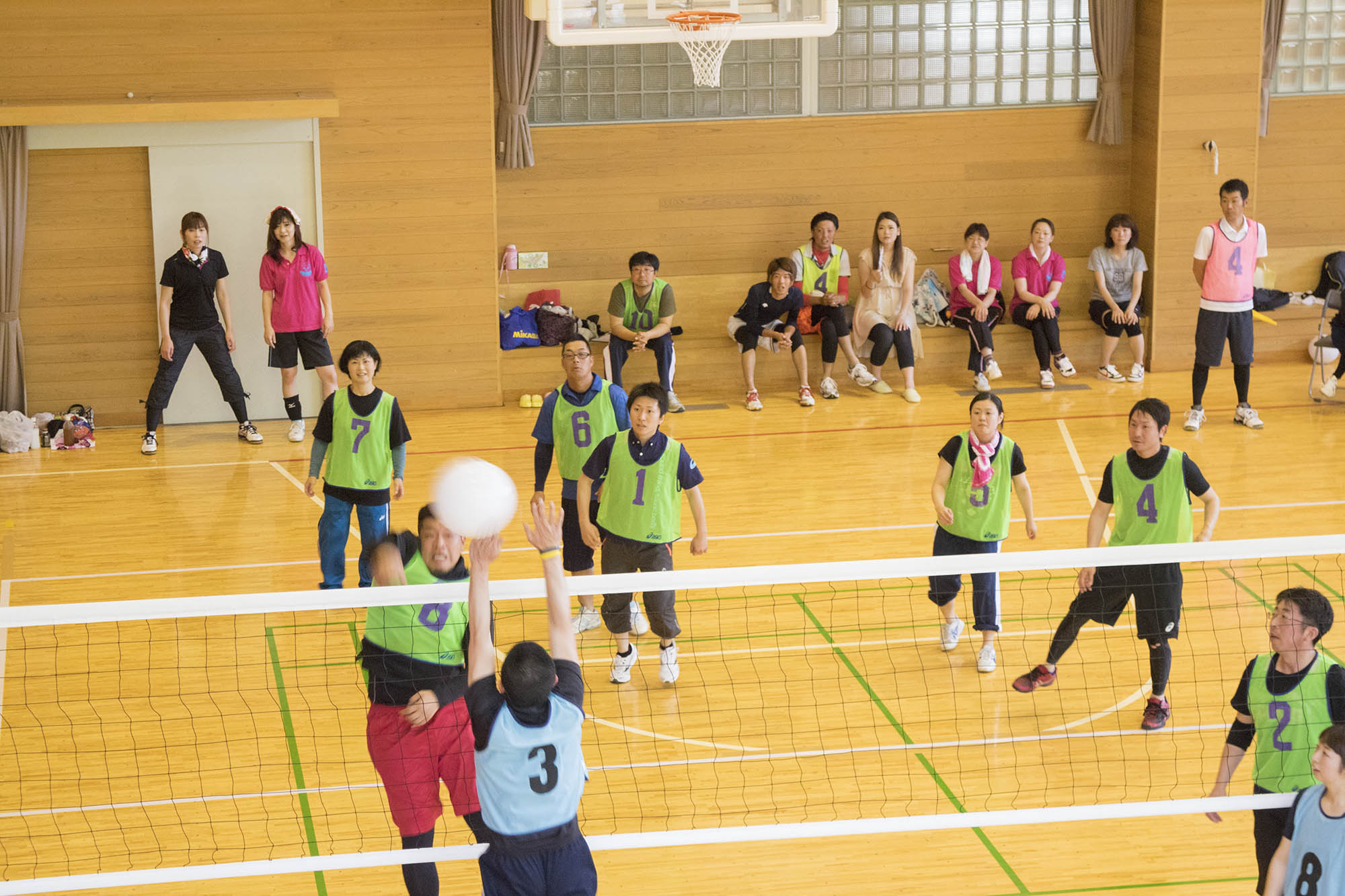 バレーボールの試合中に緑のビブスの選手がアタックをし青いビブスの選手がジャンプしてをブロックしており、緑のビブスの選手たちが真剣な表情でプレーを見守る試合中の写真