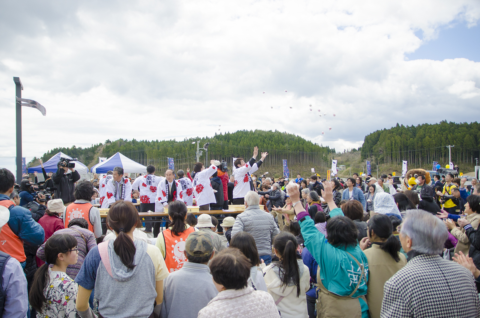 広い会場に設置された舞台の上に白い法被を着た関係者の方々が立ち、周りに集まった大勢の観客に向けて餅まきをしている写真