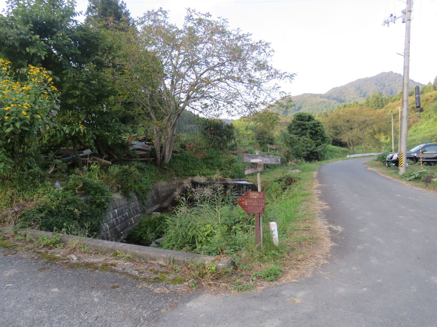 小さな側溝や水路があり、古びた看板と錆びた金属製の矢印看板が立っている写真