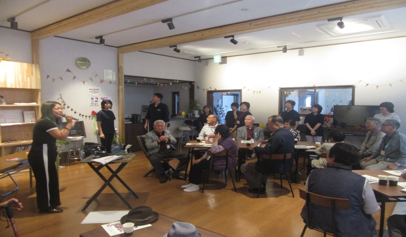 参加者と見学者を含めた十数名の高齢者が会場に集まり、司会の女性がマイクを使って話している中で皆が楽しそうに注目して見ている写真