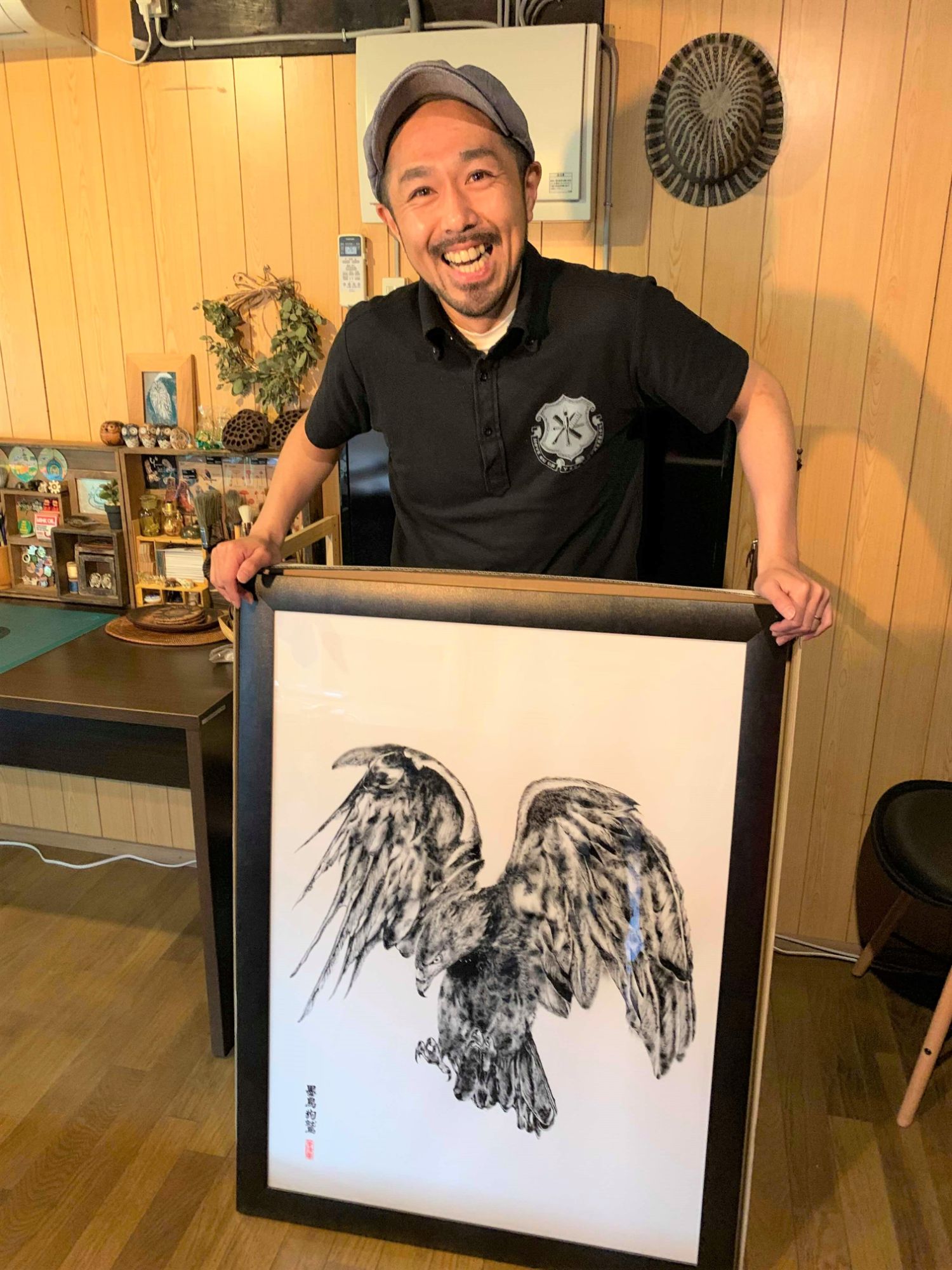笑顔の男性が室内で翼を広げた大きな黒いイヌワシを描いた額入りの作品を両手で持って誇らしげに見せている様子の写真
