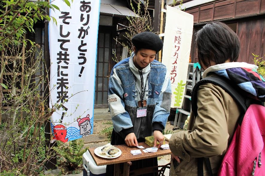 「三陸ワカメまつり」と書かれている旗が立つ茶色い外壁の家の前で、デニムジャケットを着た女性が貝殻などを並べた机の前に立ち女性客と話している写真