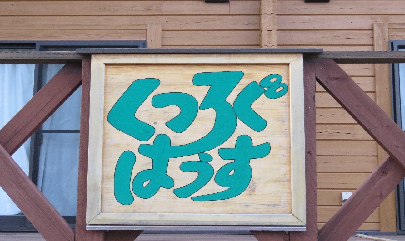 ログハウス『くつろぐはうす』の木製のフェンスに設置されている「くつろぐはうす」と緑色の文字で書かれた木製の看板をアップで写した写真