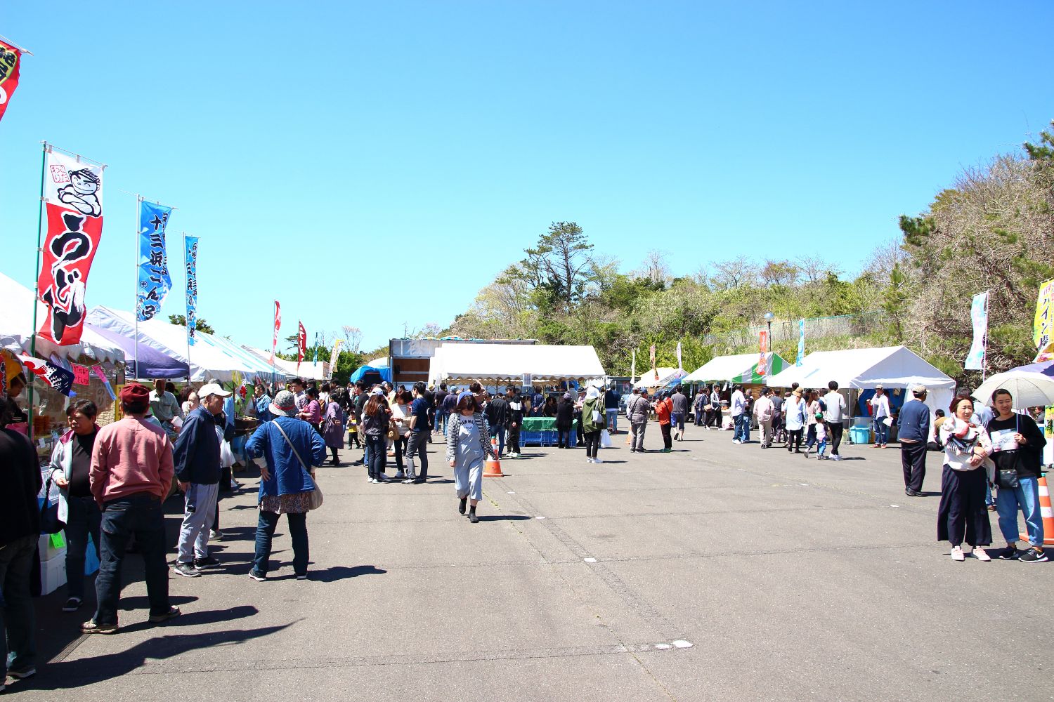 空は快晴で木々に囲まれた広々とした祭り会場の両脇にたくさんのテントが立ち並び、そこに集まった人々が買い物や食事を楽しんでいる祭りの様子の写真
