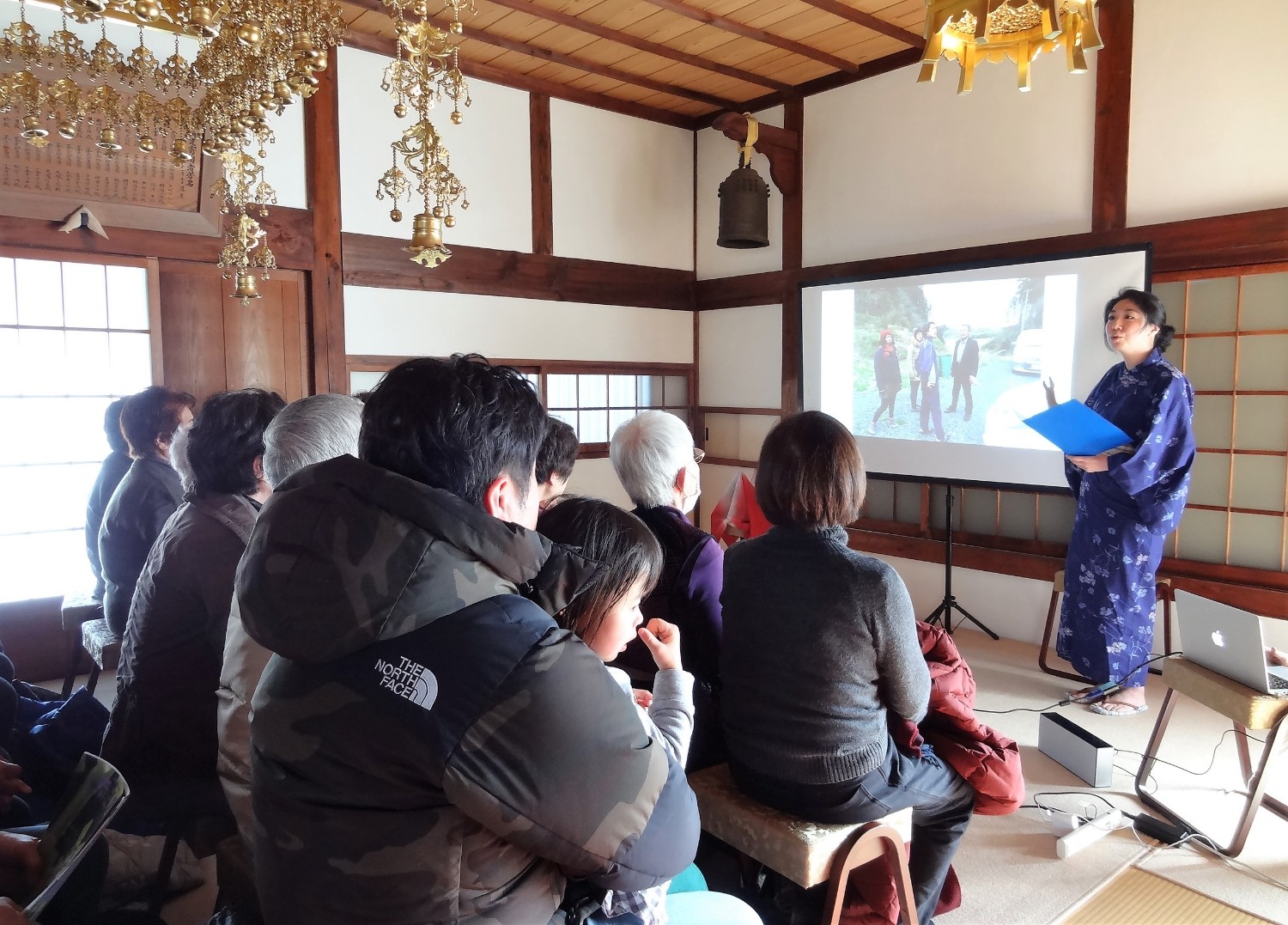 畳敷きの和室で数人が座ってスクリーンに映し出された写真を見つめながら、着物を着た人物の説明に耳を傾けている写真