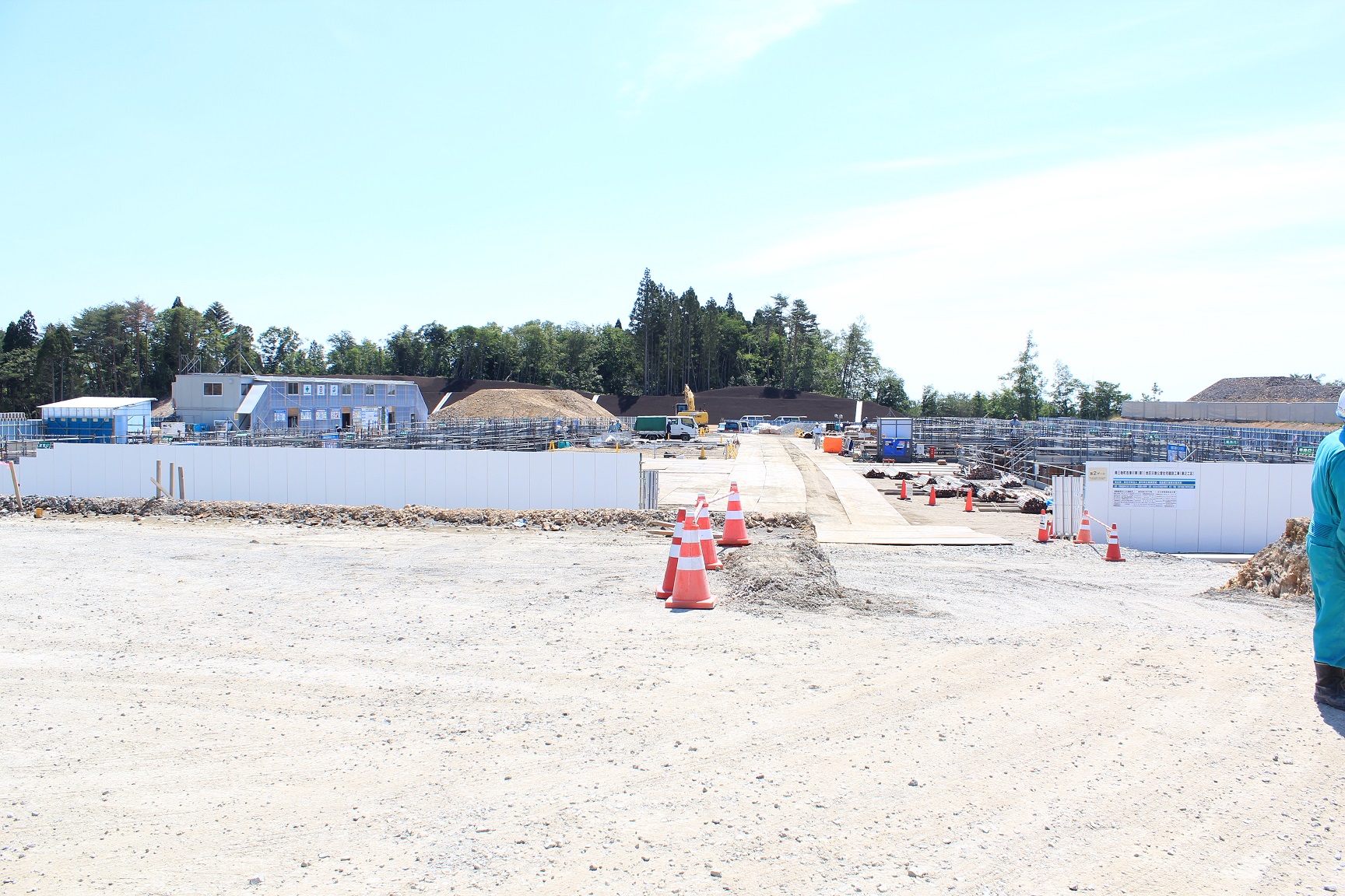 中央に工事用の道路があり、その両側には工事用の資材や機械が並んでおり、背景には木々が見える建設現場の写真