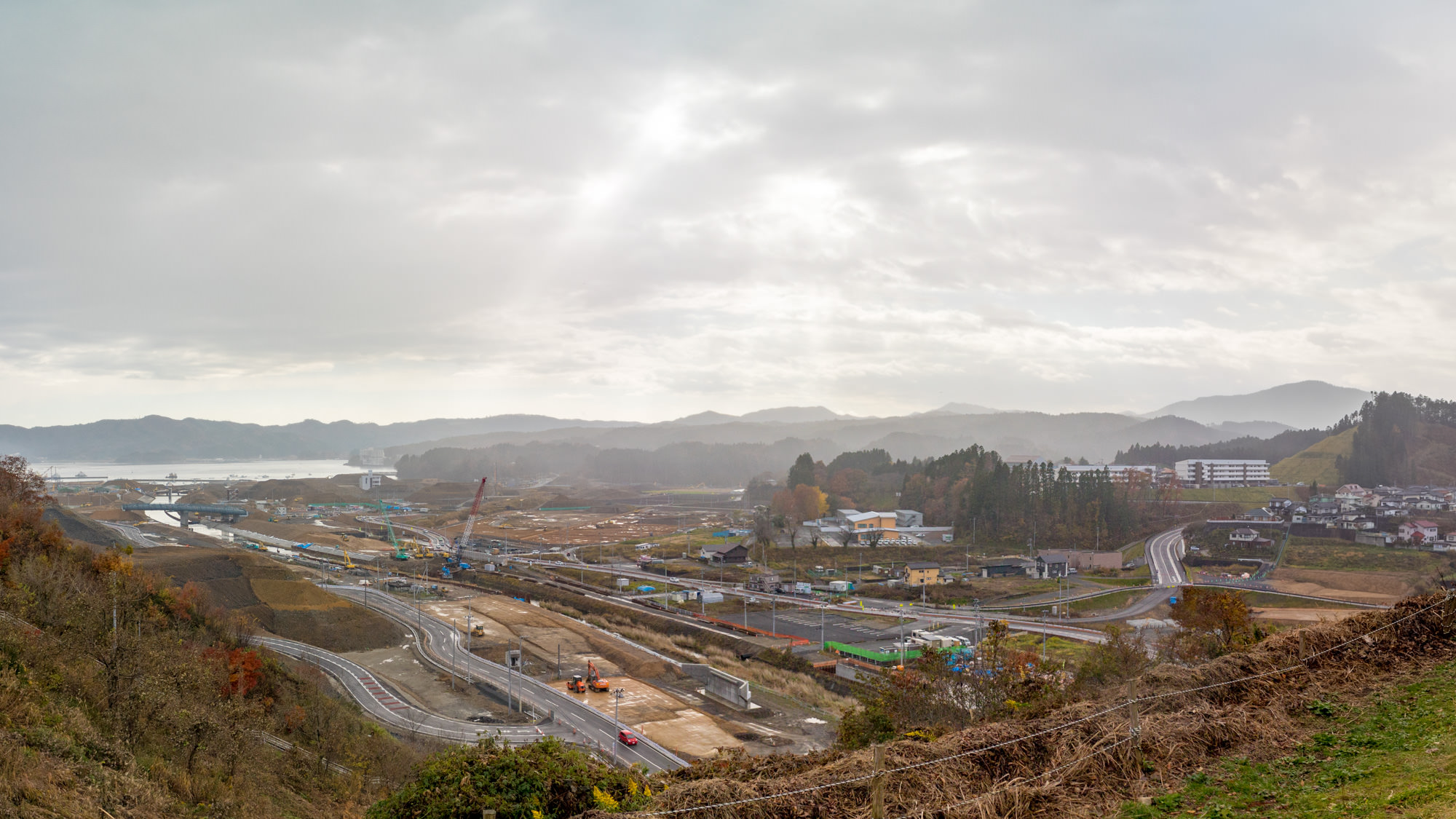 複数の道路があり、一部は工事中で、建設車両や機材が点在し、周囲には丘や木々、点在する建物が見られ、背景には山々が広がっており空は雲が広がって曇っている進行中の建設作業現場を写した写真
