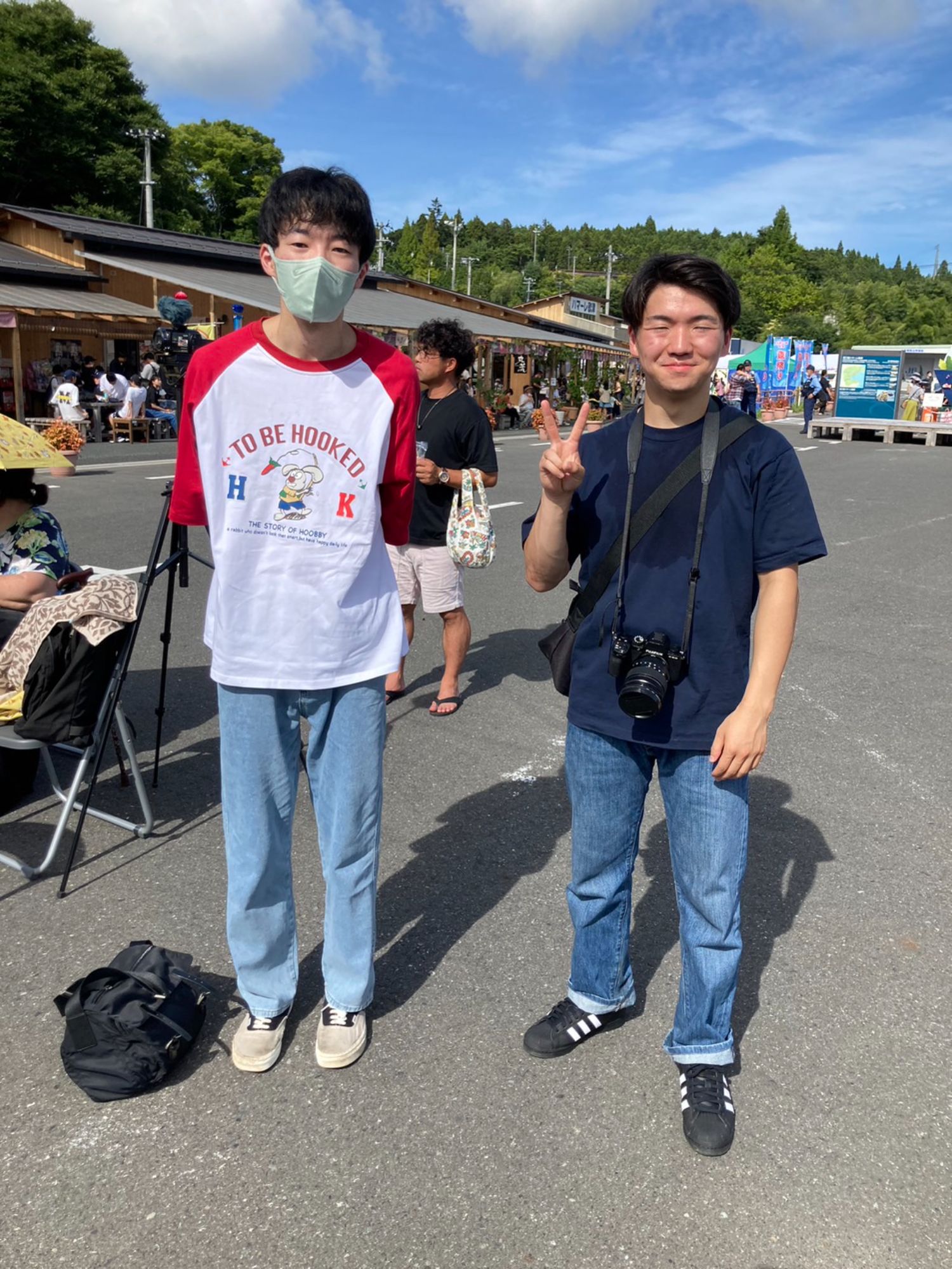 青空の下でマスクを着けた平野くんとカメラを持った加藤くんが夏祭りの会場でピースサインをしている写真