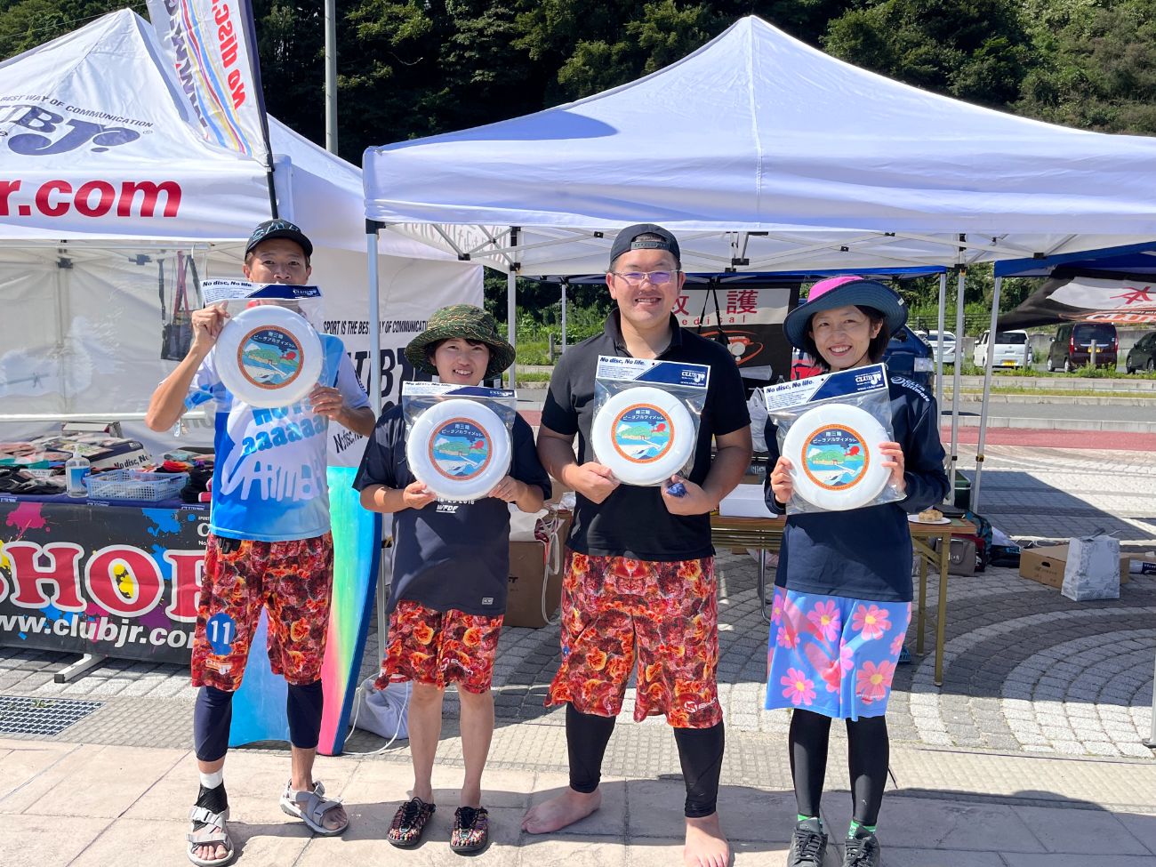 白いテントの前で、4人の男女が屋外でオリジナルディスクを手に持ち、笑顔でカメラに向かって立っている写真