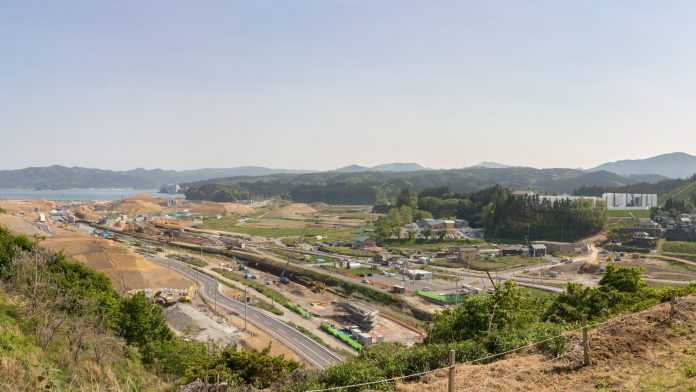 山あいの広大な地域でインフラ工事が進む様子を捉えた定点カメラによる昼間の風景で、クレーンや重機が点在し、整備中の道路や造成地が広がり、奥には海と山並みが見える写真