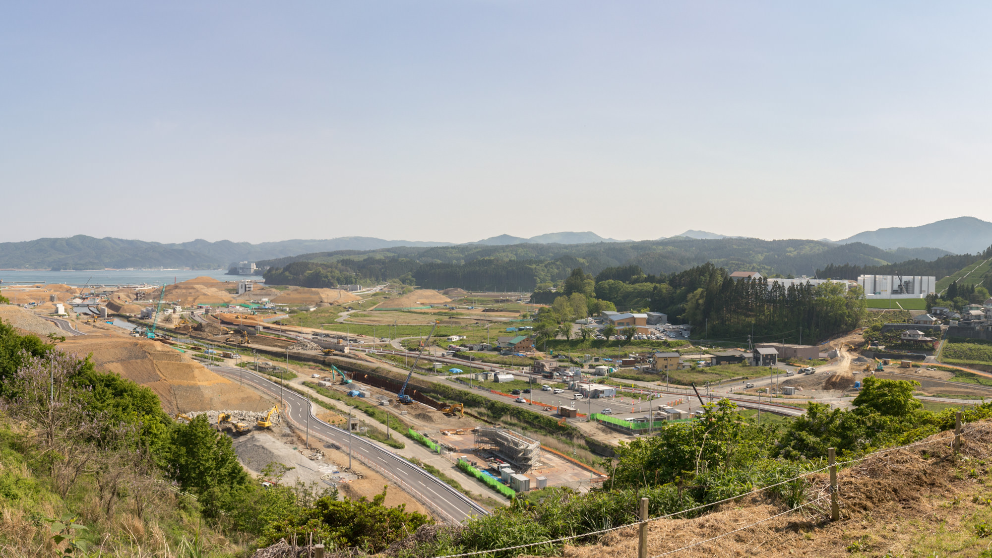 見晴らしの良い高台から見下ろす定点カメラによる昼間の風景で手前には緑の草地と土の斜面があり、その下には道路や工事現場が広がっておりショベルカーやクレーンなどの大型重機が点在し、複数の建設作業が同時進行している様子を写した写真