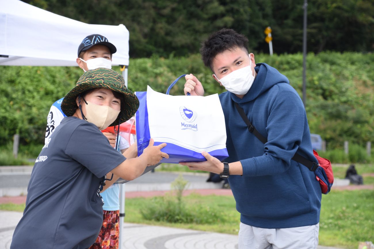 屋外のテント前でマスクを着用した男女が、大きな白と青の手提げ袋を両手で受け渡している写真